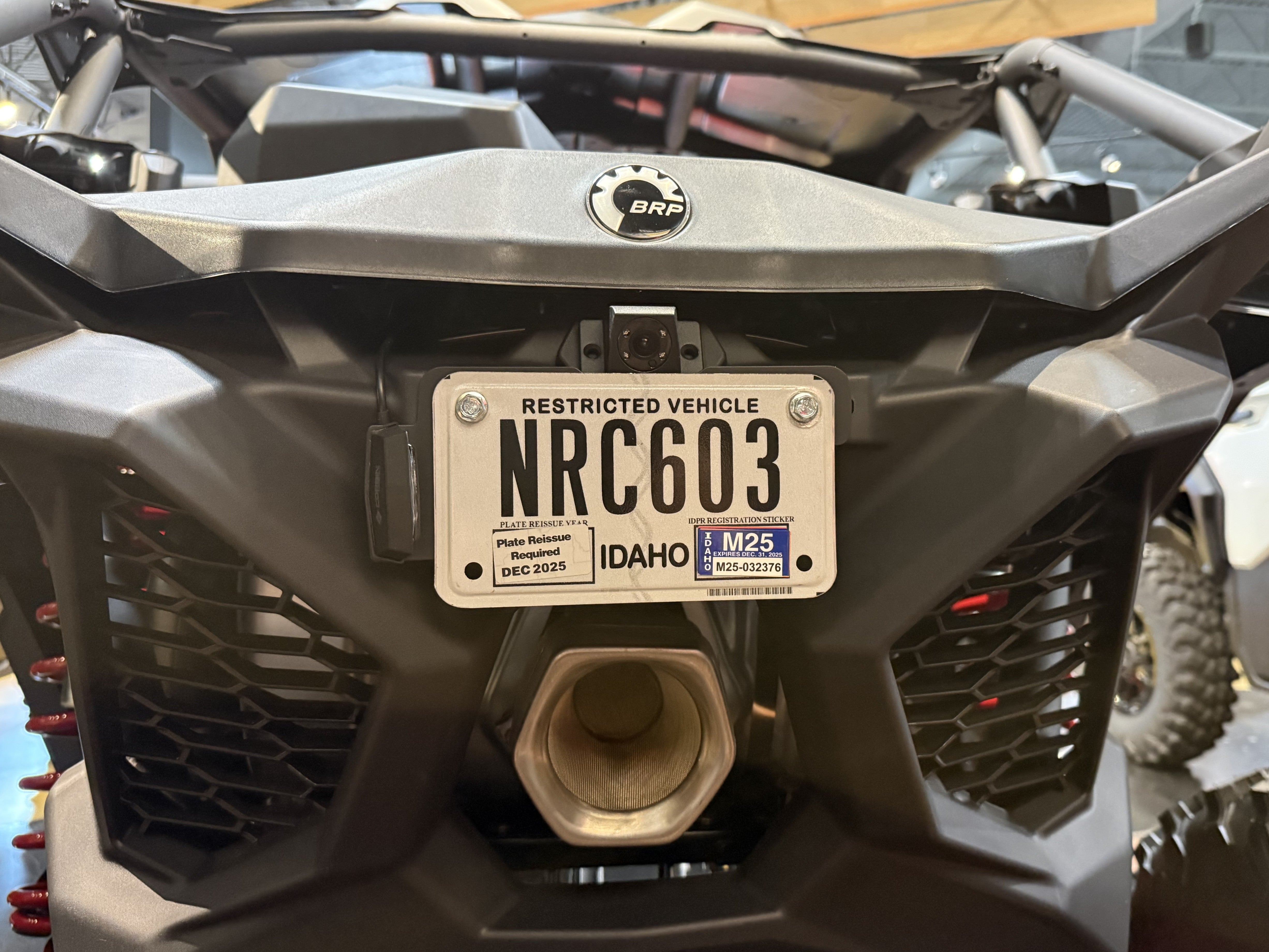 Close-up of the front of a gray and black Can-Am all-terrain vehicle, showing the, exhaust, Corbin Custom Works license plate bracket with light, and a visible BRP logo badge.
